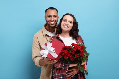 Photo of Lovely couple with gift box and bouquet of red roses on light blue background. Valentine's day celebration