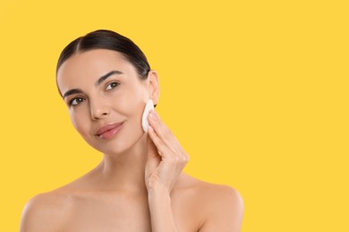 Beautiful woman removing makeup with cotton pad on yellow background. Space for text