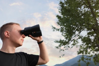 Young man with binoculars in mountains, space for text