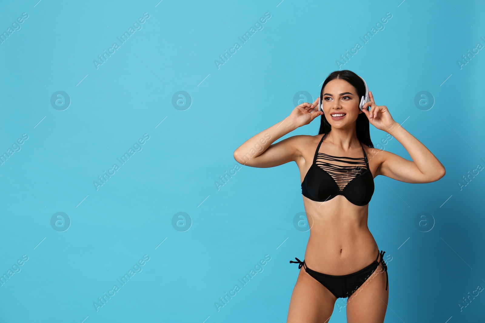 Photo of Beautiful young woman in black bikini with headphones on light blue background. Space for text