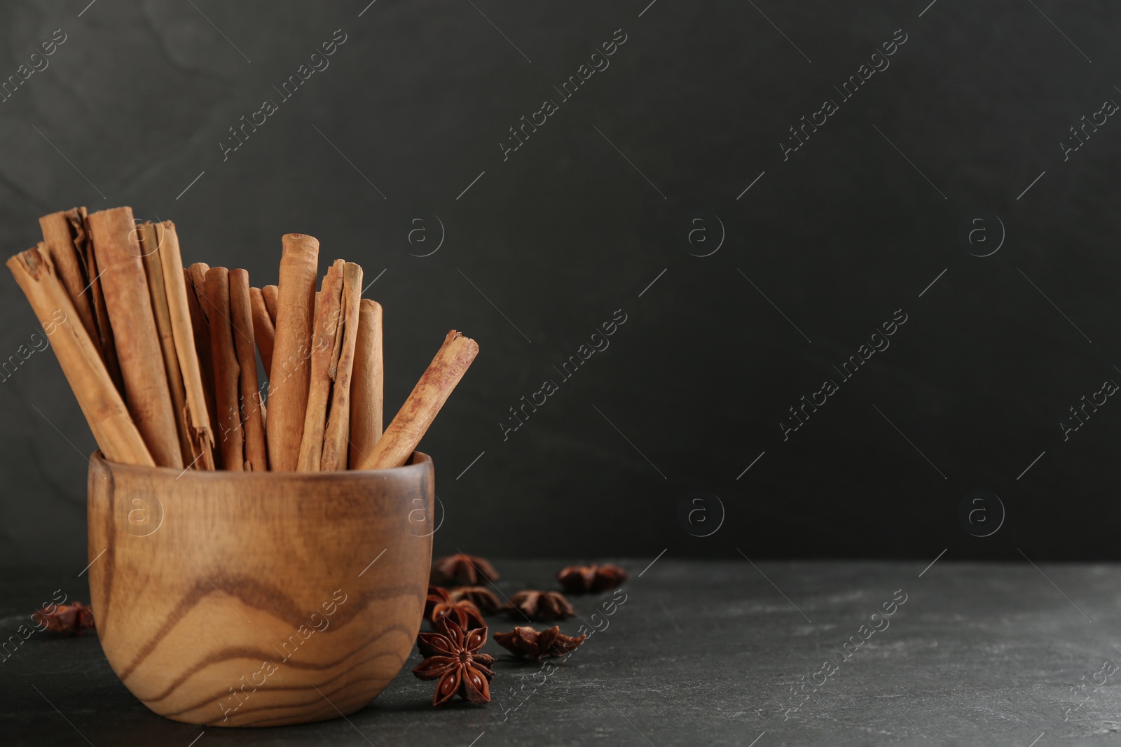 Photo of Aromatic cinnamon sticks in wooden bowl and anise on black table. Space for text