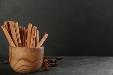 Photo of Aromatic cinnamon sticks in wooden bowl and anise on black table. Space for text