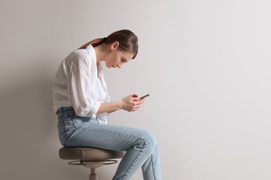 Photo of Woman with bad posture using smartphone while sitting on stool against light grey background, space for text