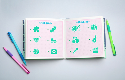 Image of Open notebook with icons of different hobbies and pens on light background, flat lay