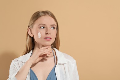 Beautiful young woman with sun protection cream on her face against beige background, space for text