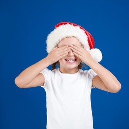 Happy little child in Santa hat closing eyes on blue background. Christmas celebration
