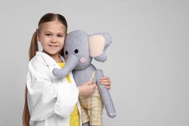 Little girl in medical uniform with toy elephant on light grey background. Space for text