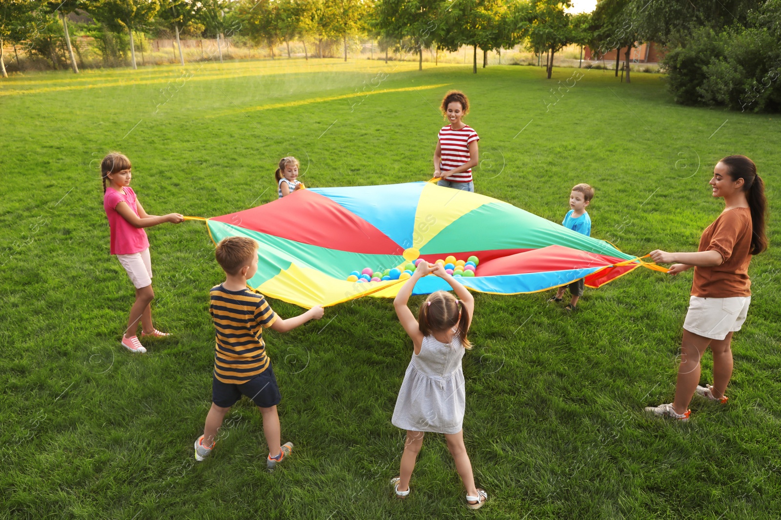 Photo of Group of children and teachers playing with rainbow playground parachute on green grass. Summer camp activity