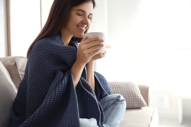 Woman covered with warm dark blue plaid enjoying hot drink on sofa at home