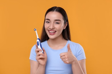 Happy young woman holding electric toothbrush and showing thumb up on yellow background