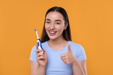 Photo of Happy young woman holding electric toothbrush and showing thumb up on yellow background