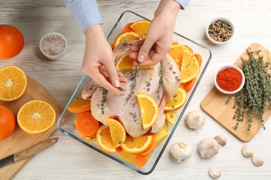 Woman adding thyme to raw chicken with orange slices at white wooden table, top view