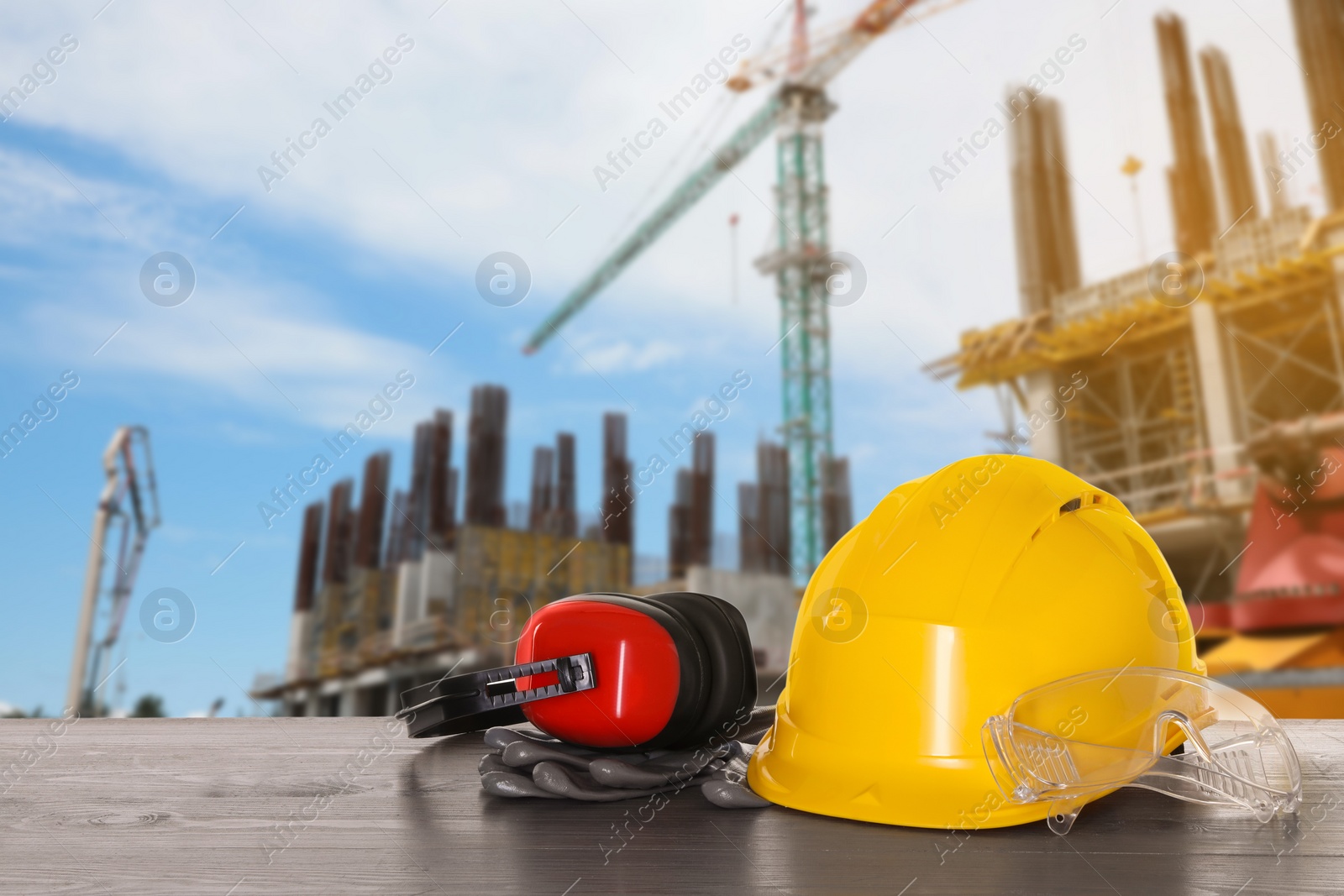 Image of Safety equipment. Hard hat, goggles, gloves and protective headphones on wooden surface near construction site with tower crane outdoors, space for text