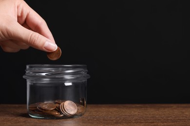 Woman putting coin in glass jar with money at wooden table, closeup. Space for text