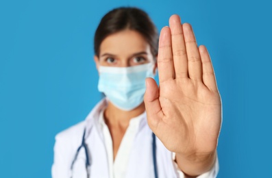 Photo of Doctor in protective mask showing stop gesture against blue background, focus on hand. Prevent spreading of coronavirus
