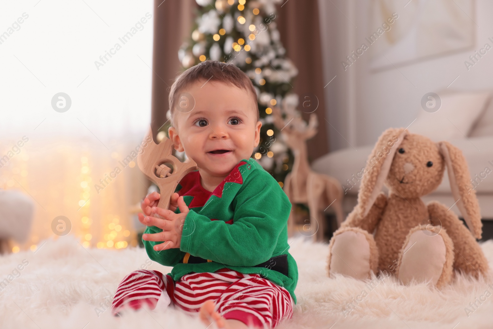 Photo of Baby wearing cute elf costume on floor in room decorated for Christmas