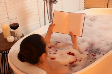 Photo of Woman reading book while taking bath in tub with foam and rose petals indoors