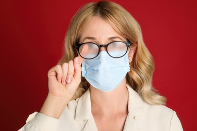 Photo of Woman wiping foggy glasses caused by wearing medical mask on red background