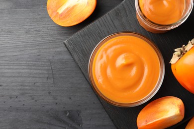 Photo of Delicious persimmon jam in glass dishware and fresh fruits on dark gray wooden table, top view. Space for text