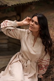 Photo of Beautiful woman wearing embroidered dress sitting near old wooden well in countryside. Ukrainian national clothes