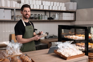 Photo of Happy seller with notebook and pen at cashier desk in bakery shop
