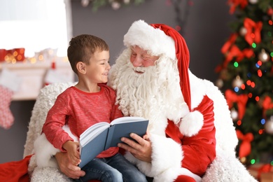 Little boy reading book while sitting on authentic Santa Claus' lap indoors