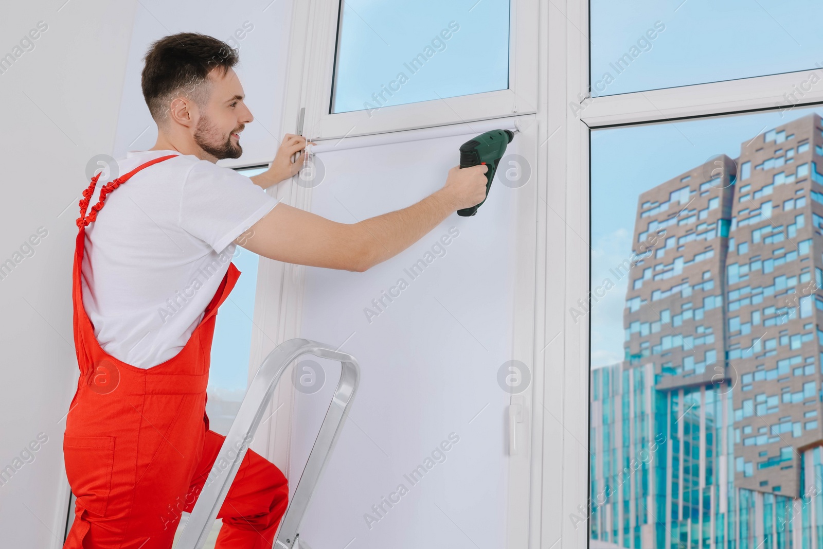 Photo of Worker in uniform installing roller window blind on stepladder indoors