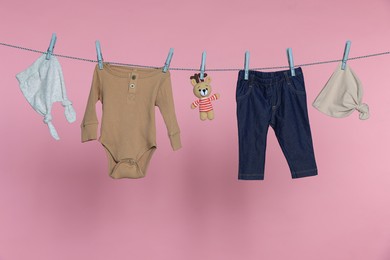 Photo of Different baby clothes and bear toy drying on laundry line against pink background