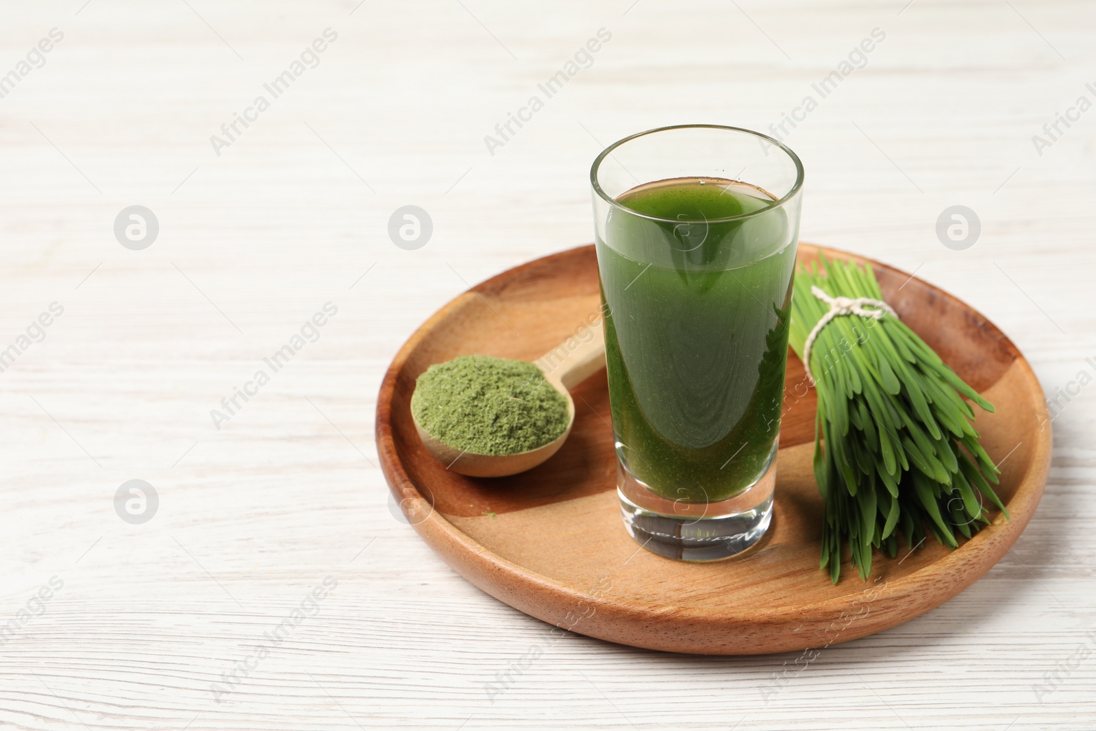 Photo of Wheat grass drink in shot glass, spoon of green powder and fresh sprouts on white wooden table, space for text