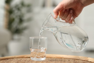 Woman pouring fresh water from jug into glass at wicker surface against blurred background, closeup