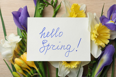 Photo of Card with words HELLO SPRING and fresh flowers on wooden table, flat lay