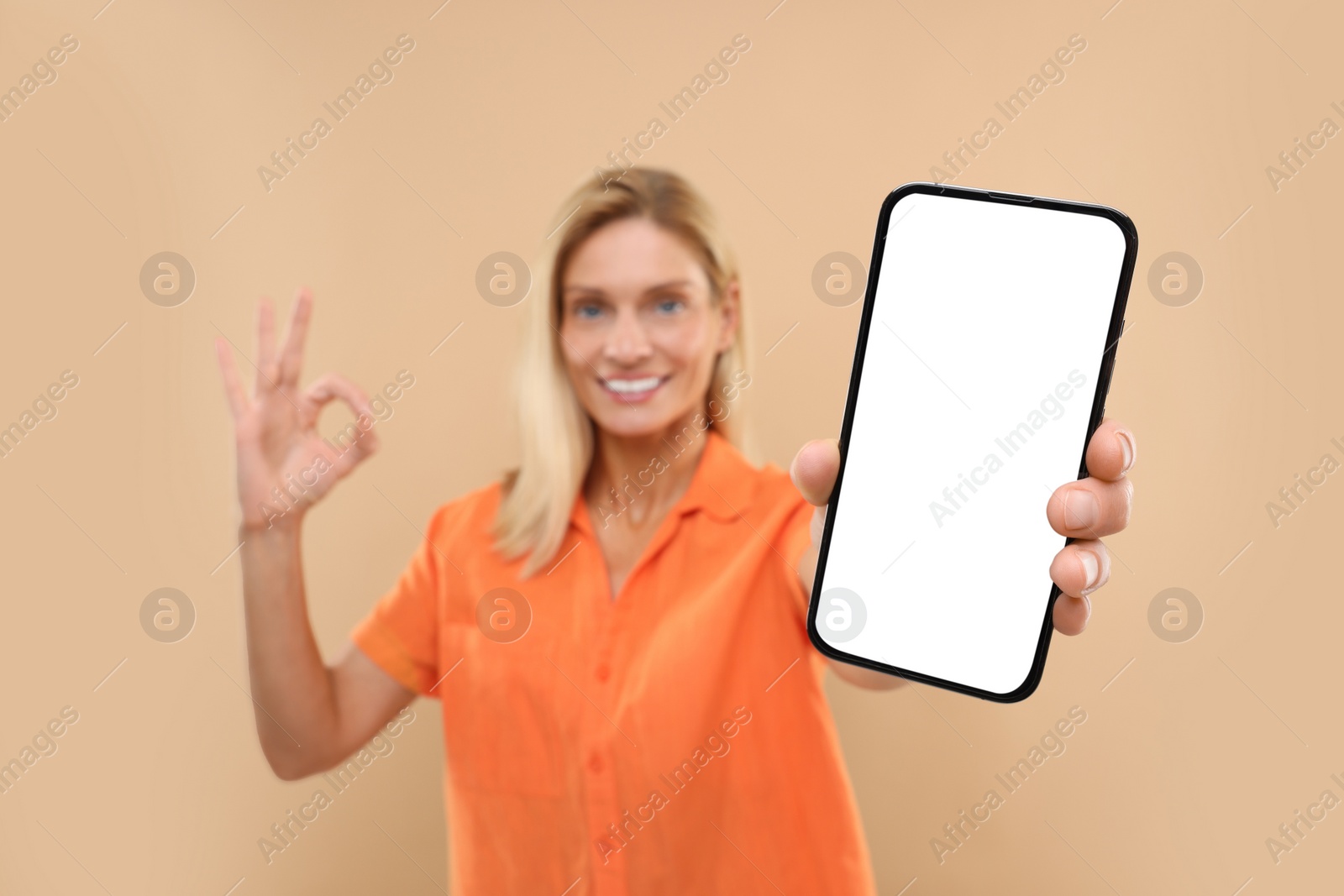 Photo of Happy woman holding smartphone with blank screen and showing OK gesture on beige background, selective focus