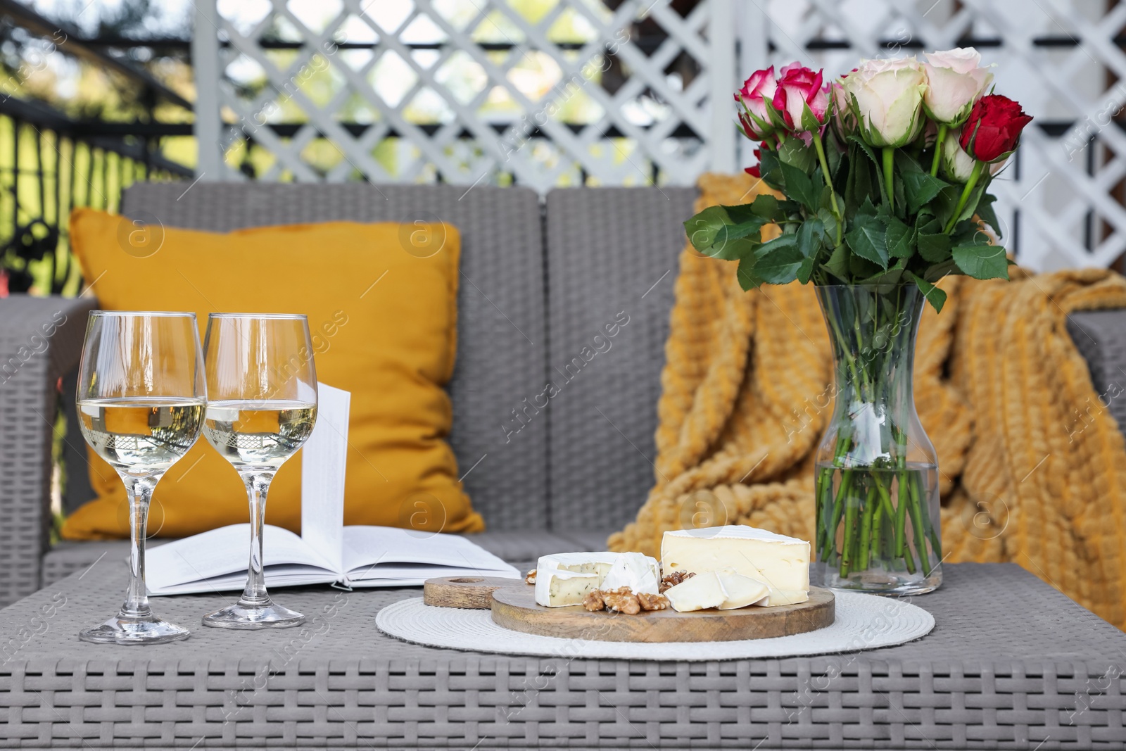 Photo of Vase with roses, open book, glasses of wine and snacks on rattan table at balcony
