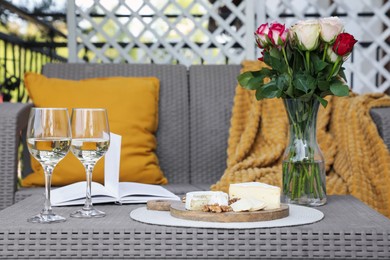 Vase with roses, open book, glasses of wine and snacks on rattan table at balcony