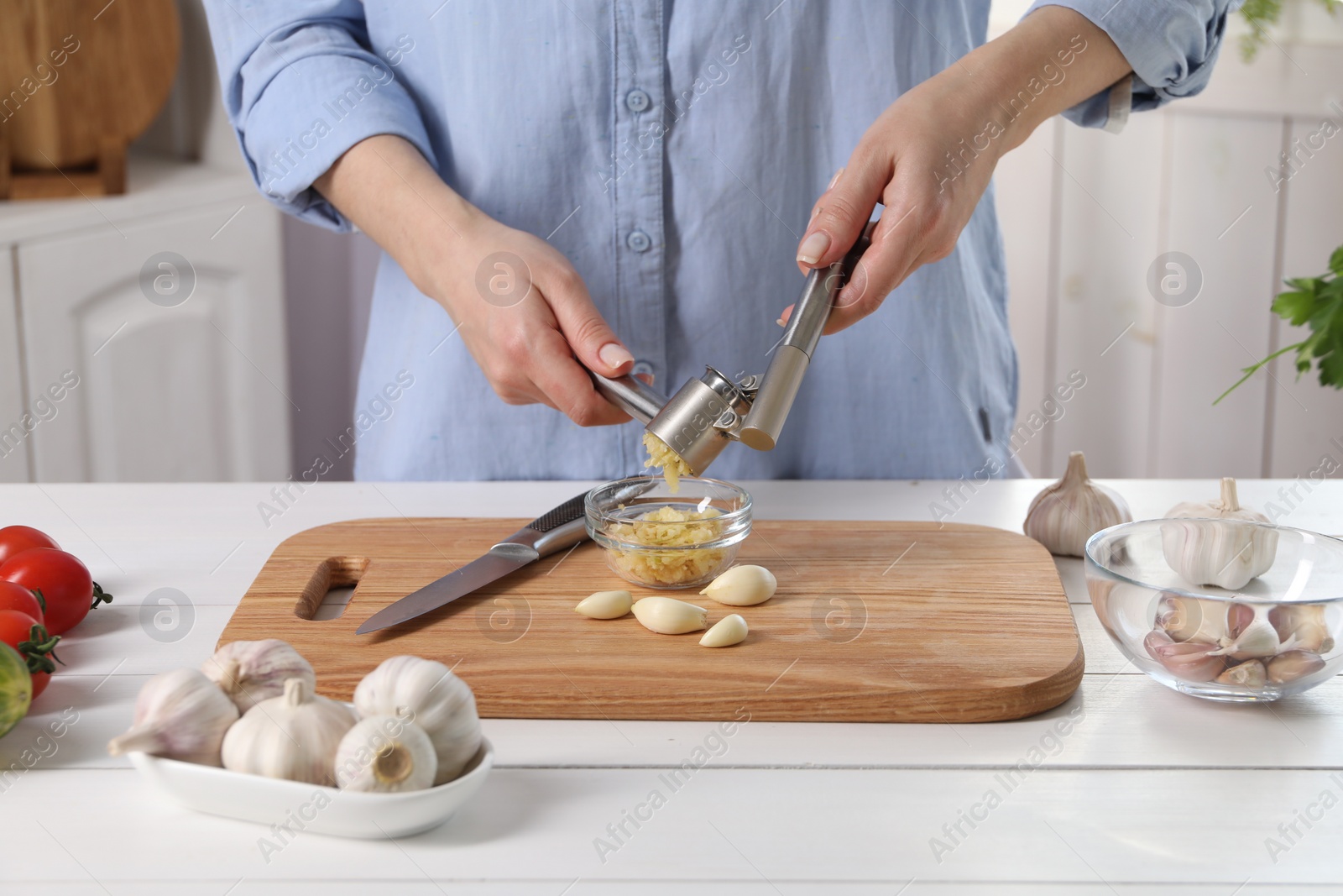 Photo of Woman squeezing garlic with press at white wooden table in kitchen, closeup