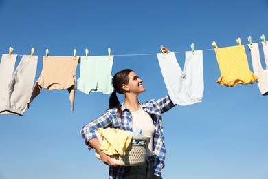 Photo of Smiling woman hanging baby clothes with clothespins on washing line for drying against blue sky outdoors