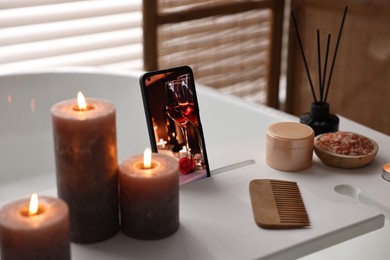 Photo of White wooden tray with smartphone, burning candles and beauty products on bathtub in bathroom