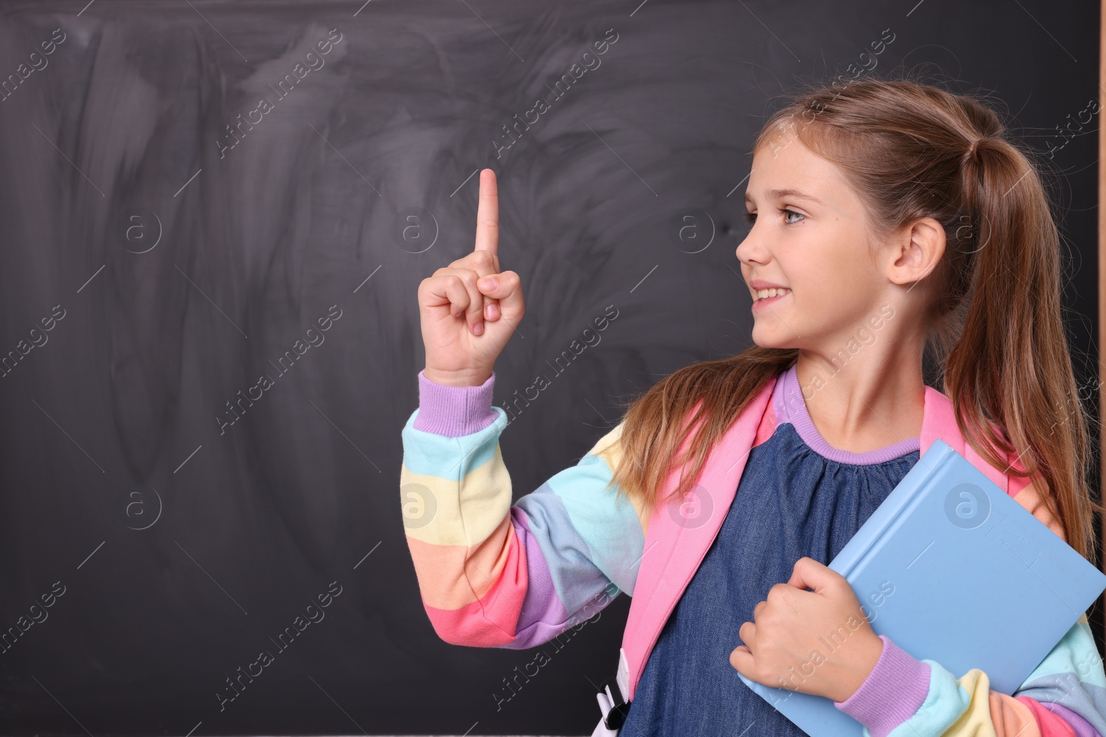 Photo of Smiling schoolgirl with book pointing at something near blackboard. Space for text