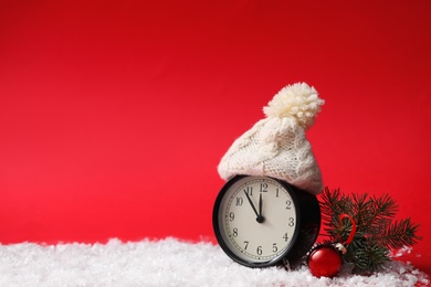 Photo of Alarm clock in hat and fir branch with Christmas ball on snow against red background, space for text. New Year countdown