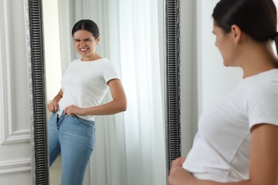 Young woman trying to put on tight jeans near mirror indoors