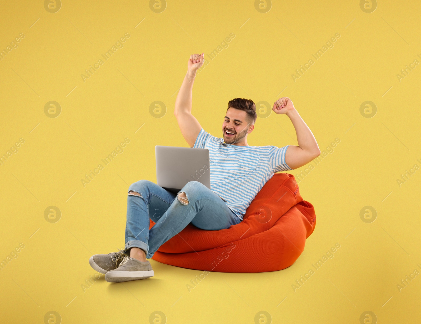 Image of Happy young man with laptop on bean bag chair, yellow background