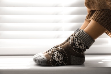 Woman wearing knitted socks on window sill indoors, closeup. Warm clothes