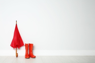 Photo of Colorful umbrella and rubber boots on floor against white wall. Space for text