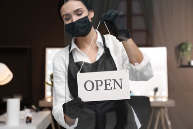 Photo of Woman hanging Open sign onto glass door in salon, focus on hands. Beauty services during Coronavirus quarantine