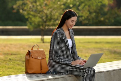Beautiful young woman with stylish backpack and laptop on bench in park