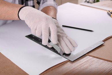 Photo of Worker cutting paper with utility knife and ruler at wooden table, closeup
