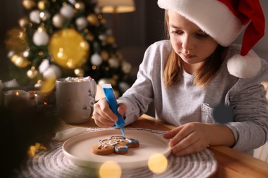 Photo of Little child in Santa hat decorating Christmas cookie at wooden table indoors