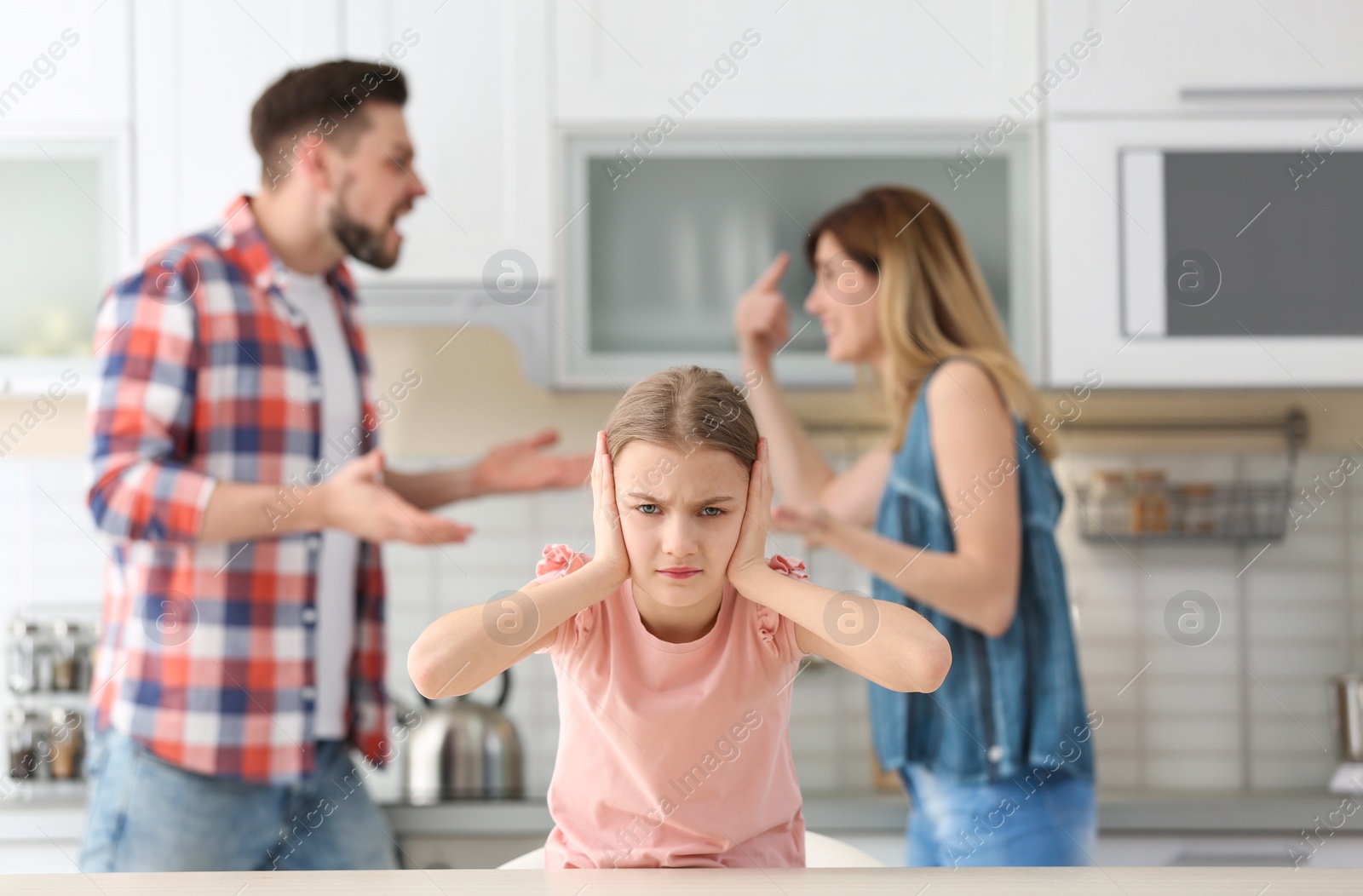 Photo of Little unhappy girl sitting at table while parents arguing on kitchen