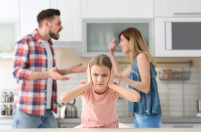 Photo of Little unhappy girl sitting at table while parents arguing on kitchen