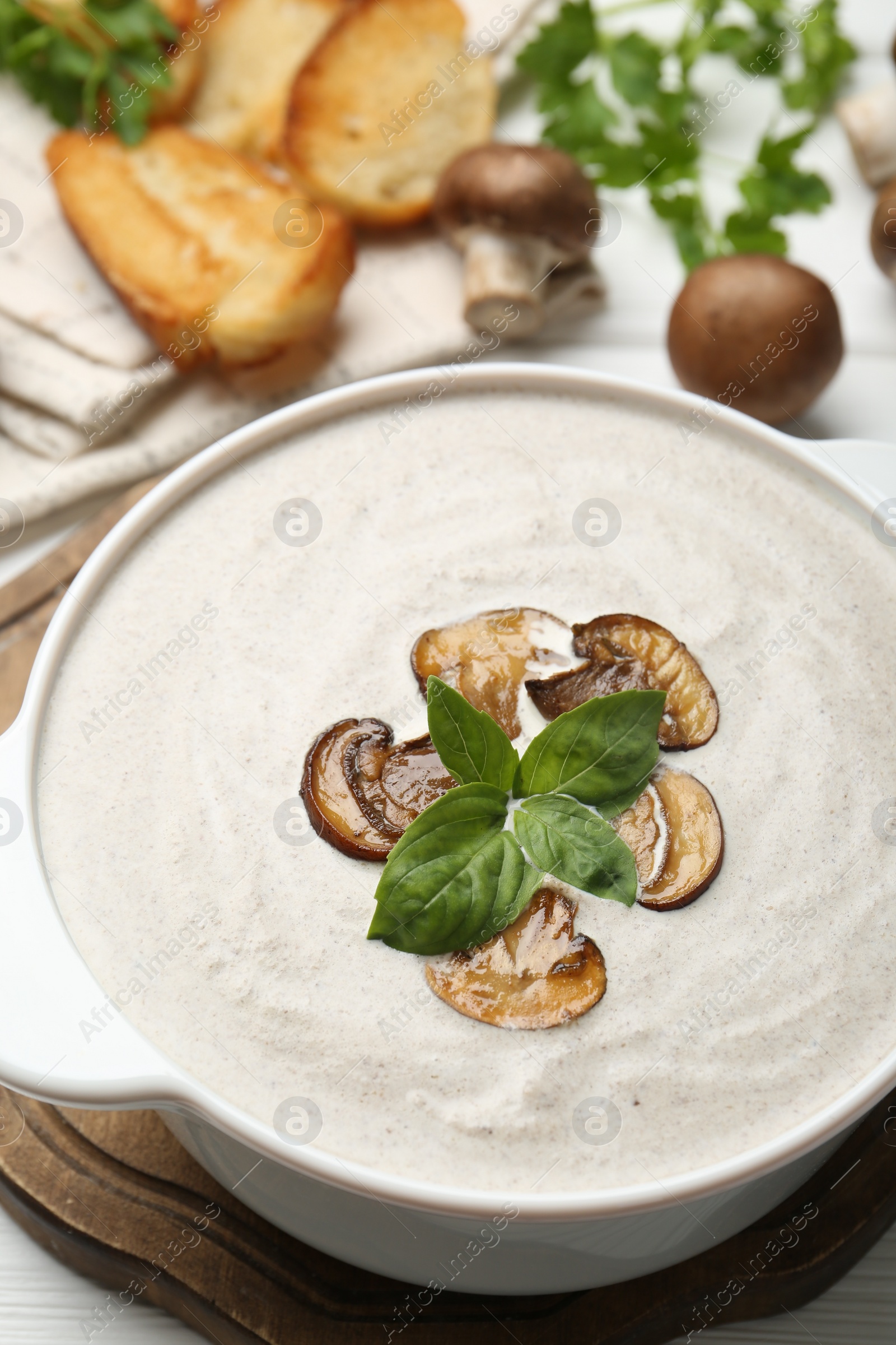 Photo of Delicious homemade mushroom soup in ceramic pot, croutons, fresh champignons and parsley on white wooden table